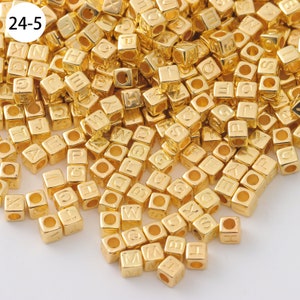 May include: A large pile of gold-colored metal letter beads. The beads are cube-shaped and have a letter embossed on each side. The beads are scattered on a white surface.