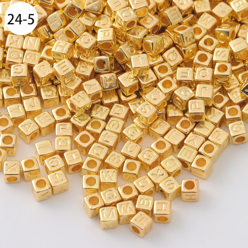 May include: A large pile of gold-colored metal letter beads. The beads are cube-shaped and have a letter embossed on each side. The beads are scattered on a white surface.