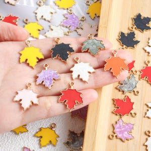 May include: A selection of colourful maple leaf charms with gold-coloured edges. The charms are in various colours, including yellow, black, white, purple, orange, red, and grey. These charms are likely used for jewellery making or other craft projects.