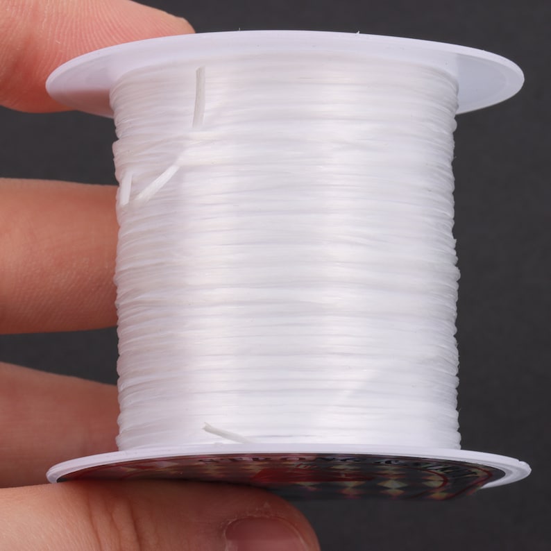 Puede incluir: Una bobina de hilo el&aacute;stico blanco, enrollada alrededor de una bobina de pl&aacute;stico blanco. El hilo se utiliza para la elaboraci&oacute;n de joyas y manualidades.