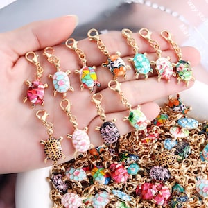 May include: A collection of gold-tone metal turtle charms, each featuring a unique colorful enamel shell pattern. The charms include lobster claw clasps for attachment. The patterns vary from leopard print and floral designs to geometric mosaics.