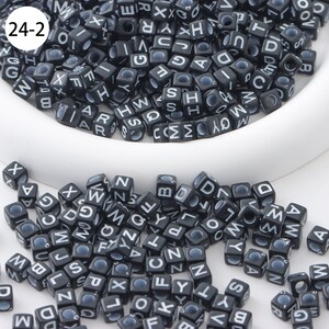 May include: A close-up of black letter beads with white lettering. The beads are scattered on a white surface. The beads are in the shape of cubes. The beads are all different letters of the alphabet.