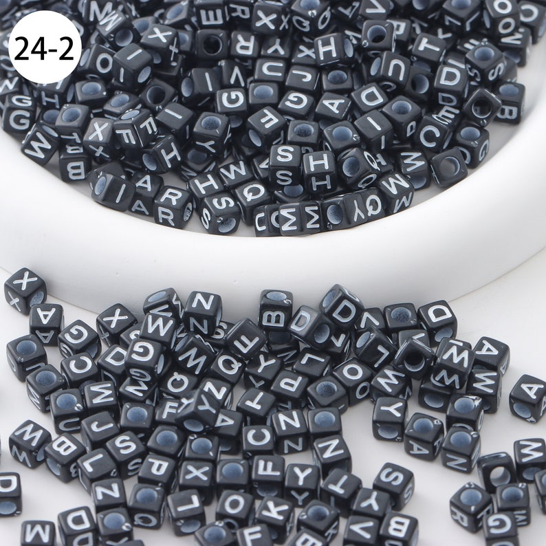 May include: A close-up of black letter beads with white lettering. The beads are scattered on a white surface. The beads are in the shape of cubes. The beads are all different letters of the alphabet.
