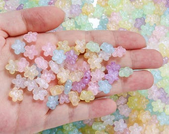 Puede incluir: Una mano sosteniendo una colección de cuentas en forma de flor de colores pastel. Las cuentas translúcidas son de tonos rosa, amarillo, azul y morado. El fondo está lleno de cuentas similares, creando una exhibición colorida y texturizada.