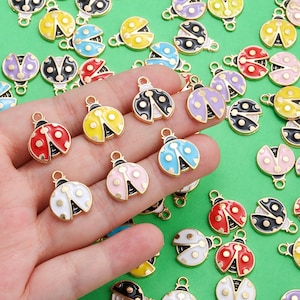 Puede incluir: Una colección de abalorios de mariquitas de colores con bordes de metal dorado. Los abalorios presentan varios colores, como rojo, amarillo, negro, blanco, rosa, azul y morado, con detalles de puntos negros. Ideales para la creación de joyas y manualidades.