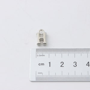 May include: Silver charm in the shape of the letter H, measuring approximately 1 cm in height. The charm has a loop at the top for attaching to a bracelet or necklace.