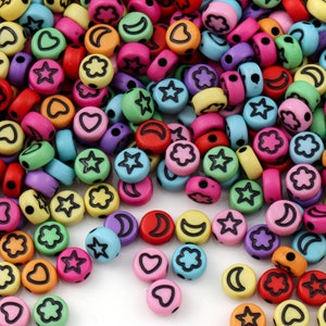 Könnte beinhalten: Eine Sammlung bunter runder Perlen mit verschiedenen schwarz umrandeten Designs, darunter Sterne, Monde, Herzen und Blumen. Die Perlen sind in den Farbtönen Rosa, Gelb, Blau, Grün, Lila und Rot gehalten, ideal für Schmuckherstellung und Bastelarbeiten.