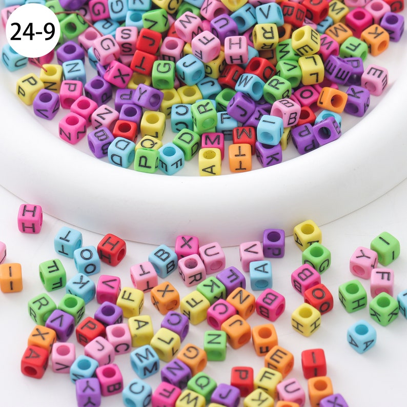 May include: A bowl filled with colorful alphabet letter beads. The beads are made of plastic and have a hole in the center. The beads are scattered on a white surface.