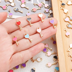 Pode incluir: Uma coleção de pequenos pingentes de chave dourados com desenhos de esmalte em forma de coração em várias cores, incluindo vermelho, rosa, azul, roxo, preto e branco. Os pingentes são exibidos em uma mão e em uma bandeja de madeira.
