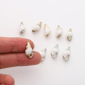 May include: A collection of ten white seashell charms with gold accents. The charms are in the shape of a seashell with a spiral design. The charms are perfect for jewellery making or crafting.