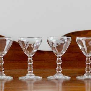 Peut inclure: Quatre verres à liqueur transparents avec des motifs de feuilles gravés. Chaque verre a une base ronde, une tige perlée et un bol évasé. Les verres sont disposés en rangée sur une surface en bois, avec un fond blanc.