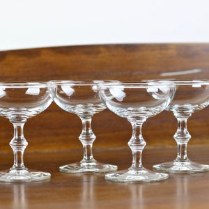 May include: Four clear champagne coupe glasses with a shallow, rounded bowl and decorative stem. The glasses are arranged on a wooden surface, with a blurred wooden background.