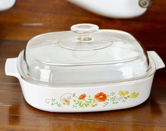 May include: White casserole dish with a clear glass lid. The dish features a floral design with red, yellow, and blue flowers. The dish has a square shape with rounded corners and handles on each side.