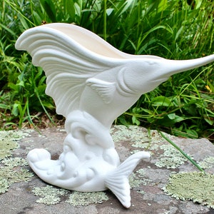 Peut inclure: Un vase en céramique blanche en forme de marlin. Le vase a un long museau pointu et un corps incurvé, avec une large ouverture en haut. Le poisson est représenté en train de sauter hors de l'eau, avec une surface texturée.