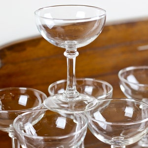 May include: A set of clear champagne coupe glasses. The glasses feature a classic design with a rounded bowl and a slender stem. The glasses are arranged in a pyramid shape, with one glass on top and the others supporting it.