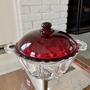 May include: A clear glass serving dish with a ruby red lid and a decorative handle. The dish has a scalloped edge and sits on a gold-colored metal stand. The lid has a round knob.