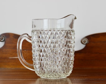 May include: Clear glass pitcher with a diamond pattern. The pitcher has a handle and a spout for pouring. The pitcher is sitting on a wooden surface. This pitcher is ideal for serving beverages.