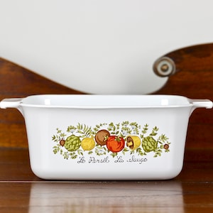 May include: White rectangular casserole dish with a colourful vegetable design. The design includes tomatoes, artichokes, and lemons. The words "Le Persil La Sauge" are printed below the design. The dish has two handles.