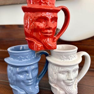 以下が含まれることがあります： 赤、青、白の3つのセラミックマグ。それぞれ顔と帽子のデザインが彫られています。赤いマグは青いマグの上に積み重ねられ、白いマグは青いマグの隣にあります。マグには側面にハンドルが付いています。