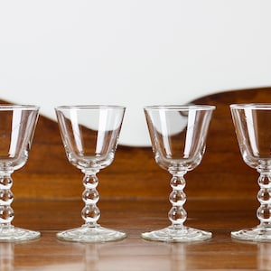May include: Four clear stemmed glasses with a rounded bowl and a beaded stem. The glasses are arranged in a row on a wooden surface, with a light-coloured background. The glasses are likely for serving wine or other beverages.
