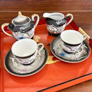 May include: A black and white porcelain tea set with dragon motifs, including a teapot, creamer, and two teacups with saucers. The set has gold trim and is displayed on a red tray.