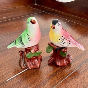May include: A pair of ceramic bird figurines. One bird is green and pink, the other is yellow and pink. Both birds perch on brown tree stump bases with green leaves. The birds have black eyes and are set against a wooden surface.