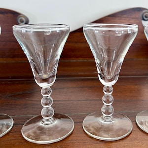 May include: Clear glass stemmed glasses with a conical bowl and a beaded stem. The glasses have a wide, flat base and are arranged on a dark wooden surface. The glasses are empty and appear to be for serving beverages.