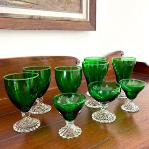 May include: A collection of emerald green glassware, including wine glasses and small stemmed glasses, arranged on a wooden surface. The glasses have clear, textured bases and a vibrant, glossy finish. The arrangement suggests a vintage or antique aesthetic.