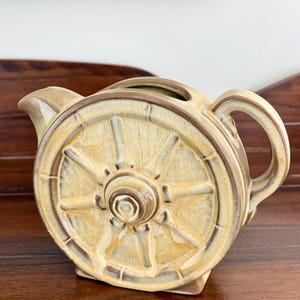 May include: A ceramic teapot with a wagon wheel design. The teapot is tan and brown with a spout, handle, and a lid opening. The wheel design is on both sides of the teapot.