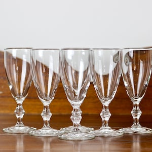 May include: Five clear glass stemmed glasses are displayed. Each glass features a fluted bowl, an ornate stem, and a circular base. The glasses are arranged in a row on a wooden surface.