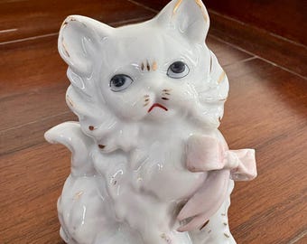 May include: A white ceramic cat figurine with gold accents on the ears and a pink bow. The cat has blue eyes and a slightly frowning expression. The figurine is a decorative item.