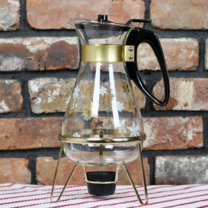 May include: A vintage glass coffee carafe with a black handle and lid, decorated with gold and white snowflake patterns. It has a gold band and is set in a gold-coloured metal stand with a black base for a warming candle. The carafe is approximately 25 cm tall.