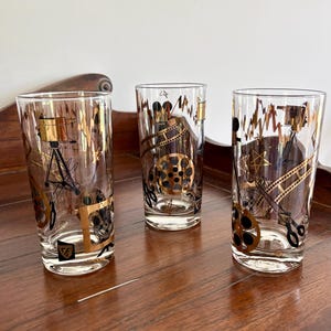 May include: Three clear drinking glasses with gold and black film reel and camera designs. The glasses are tall and cylindrical, with a gold rim and base. The film reel and camera designs are repeated on each glass.