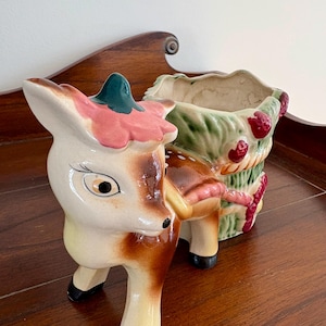 May include: A vintage ceramic planter in the shape of a deer pulling a basket. The deer is cream, brown, and black with a pink and green floral accent. The basket is green with red berries and rope detailing.