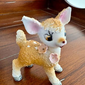 May include: A ceramic deer figurine with a textured, tan body and white accents. The deer has large, pink-lined ears, a white face, and a red nose. Two pink flower embellishments adorn its side. The figurine stands on four legs with black and white hooves.