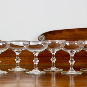 May include: A set of six clear champagne coupe glasses. Each glass has a shallow, rounded bowl, a decorative stem, and a circular base. The glasses are arranged in a row, reflecting light, against a wooden background.