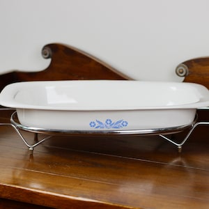May include: A white oval casserole dish with blue floral detailing, on a chrome metal stand with wooden handles. The dish is for serving food and is on a wooden surface. The dish's dimensions are approximately 30 cm.