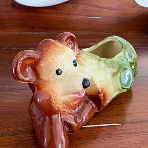May include: A ceramic bear-shaped planter. The bear is painted in shades of brown and tan, with black eyes and nose. The planter section is green and tan. The bear is in a reclining pose.