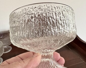 May include: Clear glass dessert cup with a textured, bark-like design. The cup has a wide, rounded bowl and a short, pedestal base. The glass is transparent, allowing light to pass through. The cup is held by a hand.