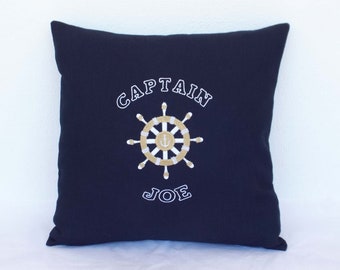 Könnte beinhalten: Ein marineblaues Kissen mit weißer Stickerei, auf dem "CAPTAIN JOE" steht und ein gold-weißes Schiffssteuerrad mit Anker in der Mitte abgebildet ist.