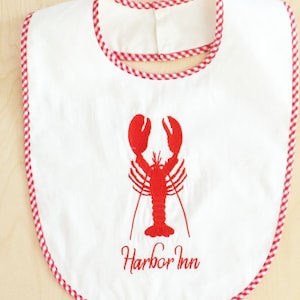 May include: A white bib featuring a red embroidered lobster centred on the front. Below the lobster, the text Harbor Inn is embroidered in red. The edges of the bib are finished with a red and white gingham check trim around the entire perimeter.