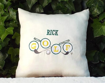 Könnte beinhalten: Ein weißes Kissen mit grüner und gelber Stickerei des Wortes "GOLF" mit Golfschlägern und Golfbällen. Der Name "RICK" ist über dem Wort "GOLF" gestickt.