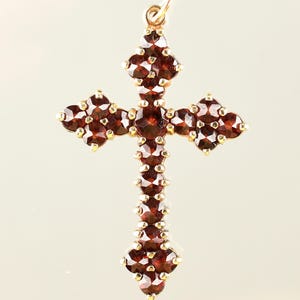 Può includere: Un pendente color oro a forma di croce, ornato da numerose piccole pietre preziose rosso scuro. Le pietre sono incastonate vicine, creando un aspetto ricco e strutturato. La croce ha un anello nella parte superiore per essere appesa.