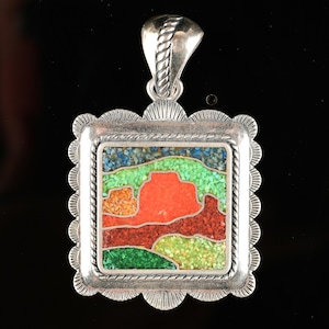 May include: A silver pendant with a square, mosaic-style design depicting a landscape scene. The landscape features red, orange, green, and blue elements, set within a scalloped frame. The pendant has a decorative bail for a chain.