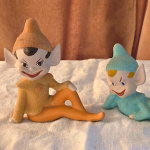 May include: Two ceramic elf figurines. One elf is wearing a yellow top and orange trousers, the other is wearing a blue top and trousers. Both have pointed ears, mischievous smiles, and are lying down.