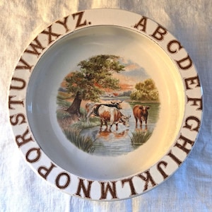 May include: A white ceramic bowl with a brown alphabet border. The central image shows cows in a river, trees, and a sunset. The letters are in a serif font. A decorative item for the home.
