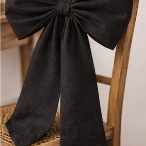 May include: A large black fabric bow is tied to the back of a wooden chair. The bow has a full shape with long, flowing tails. The chair is made of light-coloured wood.