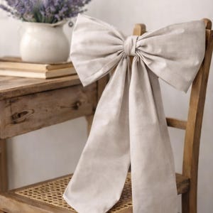 May include: A large, beige fabric bow tied to the backrest of a wooden chair with a woven seat. The bow features two wide, long tails that drape downward from the centre knot, made of a solid, light-coloured textile material with a visible weave.