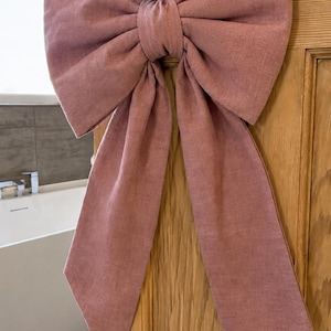 May include: A large, dusty rose-coloured fabric bow with two long, hanging tails. The bow is tied securely around a round, brass-coloured metal door knob attached to a wooden door with a visible grain pattern and a rectangular brass backplate.