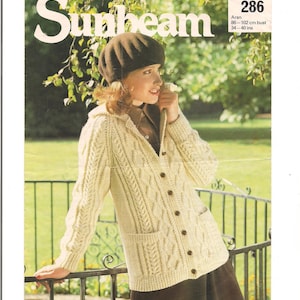 May include: A white knitted cardigan with a cable knit pattern and a brown button closure. The cardigan has two pockets and is worn by a model with a brown beret. The image includes the text "Sunbeam" and "286 Aran 86-102 cm bust 34-40 ins"